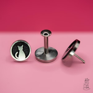 以下が含まれることがあります： ピンク色の背景に、3つの銀色の猫をテーマにしたジュエリー。 黒い背景と白い猫のシルエットが付いた2つの丸いスタッドと、バーベルスタイルのピースが含まれています。