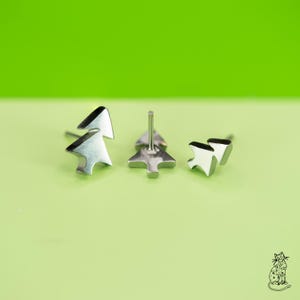 May include: Three silver-colored tree-shaped stud earrings. The earrings are displayed on a light green surface against a bright green background. The earrings are made of a shiny metal and have a simple, minimalist design. The earrings are a festive accessory.