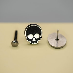 May include: A set of three metal earrings. One is a post earring with a round silver ball, another is a skull-shaped stud with a black background and white skull design, and the third is a silver disc with a post.