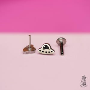 Puede incluir: Tres piezas de joyería corporal plateadas. Una es una pequeña forma de corazón marrón y plateado. Otra es una pequeña forma de OVNI plateada con detalles en blanco y negro. La tercera es un poste plateado con una parte superior plana y redonda.