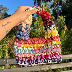 May include: A small, rectangular handbag crafted from thick, multicolored fabric yarn. The bag features a repeating horizontal striped pattern in shades of red, yellow, blue, pink, and purple, with a single integrated handle at the top.