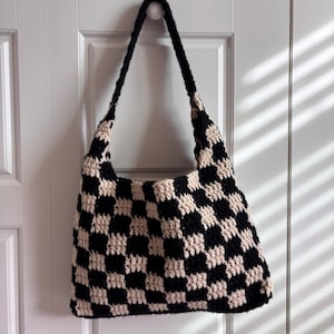 May include: A handmade crochet shoulder bag featuring a repeating black and cream checkerboard pattern. The bag has a single wide strap and a rectangular body constructed from yarn, showcasing a textured, woven stitch throughout the design.