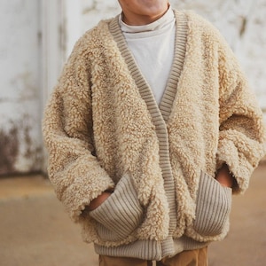 May include: A beige, textured, fleece-like cardigan featuring a V-neckline, long sleeves, and two large front pockets. The garment is accented with ribbed knit trim along the collar, cuffs, hem, and pocket openings, worn over a plain white shirt.
