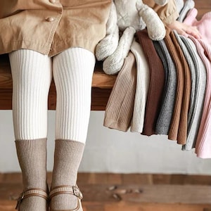 Könnte beinhalten: Ein Kind trägt cremefarbene Rippstrumpfhosen mit braunen Socken und Schnallenschuhen. Eine Auswahl an Rippstrickpullovern in Braun-, Grau- und Rosatönen wird auf einer Holzoberfläche präsentiert. Ein Stofftier ist ebenfalls vorhanden.