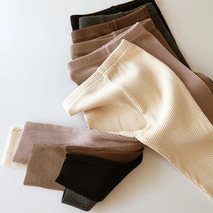 Könnte beinhalten: Eine Auswahl an gerippten Leggings in verschiedenen neutralen Farben, darunter Creme, Beige, Braun und Schwarz. Die Leggings sind ordentlich angeordnet, um ihre Textur und Farbvariationen zu zeigen. Der Stoff wirkt weich und bequem.