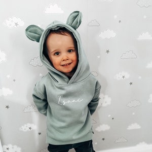 May include: A sage green hooded sweatshirt with attached ears, worn by a child. The name "Isaac" is written in cursive on the front. The child is wearing black pants. The background is a light gray wall with a cloud and star pattern.