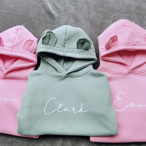 May include: Three hooded sweatshirts in pink and sage green, each with a name in white script. The hoods have small, rounded ear-like details. The names are "Maisie", "Clark", and "Evie".
