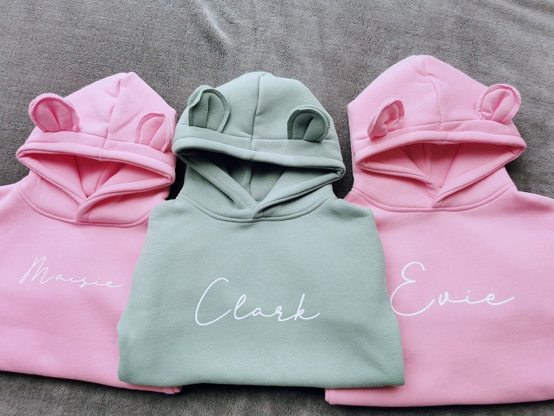 May include: Three hooded sweatshirts in pink and sage green, each with a name in white script. The hoods have small, rounded ear-like details. The names are "Maisie", "Clark", and "Evie".
