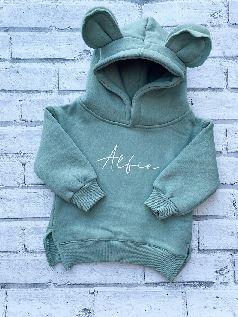 May include: A sage green baby hoodie with a hood featuring bear ears. The word "Alfie" is written in cursive on the front. The hoodie has long sleeves and a ribbed hem and cuffs. The background is a white brick wall.