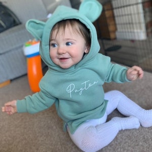 May include: A smiling baby wearing a mint green hoodie with large ears and the name "Payton" in cursive. The baby is also wearing white patterned leggings. A colorful toy is visible in the foreground.