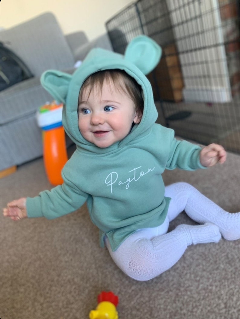 May include: A smiling baby wearing a mint green hoodie with large ears and the name "Payton" in cursive. The baby is also wearing white patterned leggings. A colorful toy is visible in the foreground.
