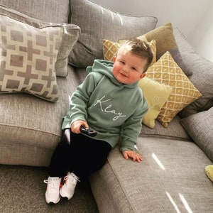 May include: A young child wearing a sage green hoodie with the name "Klay" in white script, paired with black pants and white sneakers with red accents. The child is holding a remote control and sitting on a gray sofa with decorative pillows.
