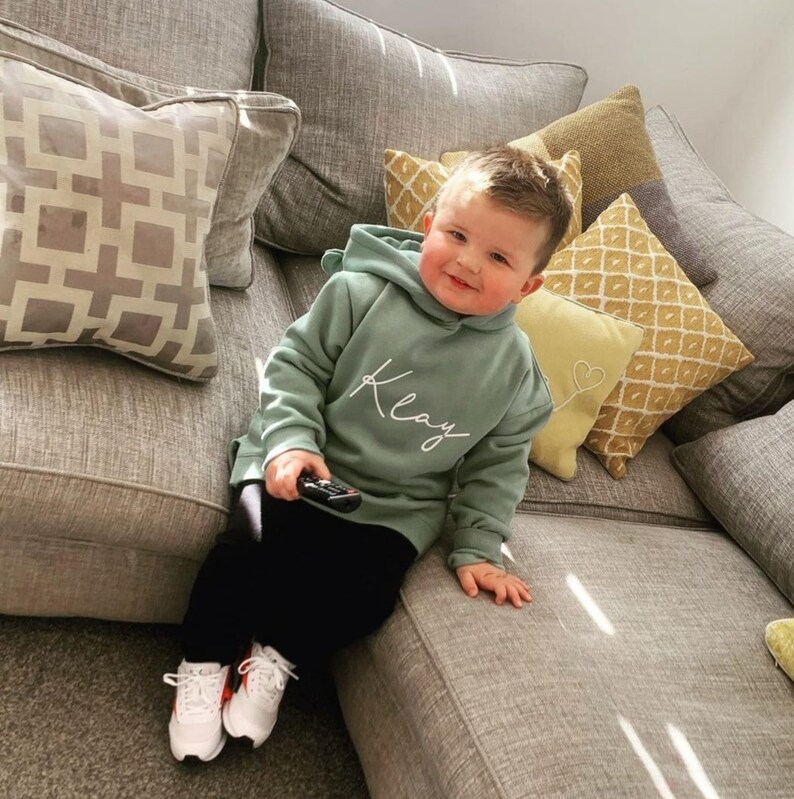 May include: A young child wearing a sage green hoodie with the name "Klay" in white script, paired with black pants and white sneakers with red accents. The child is holding a remote control and sitting on a gray sofa with decorative pillows.