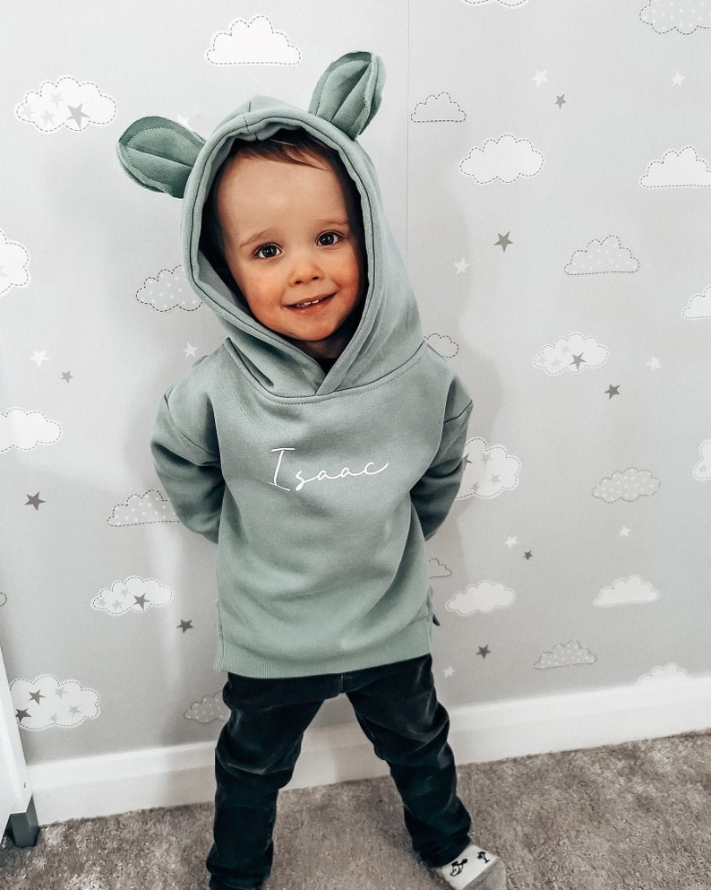 May include: A sage green hooded sweatshirt with attached ears, worn by a child. The name "Isaac" is written in cursive on the front. The child is wearing black pants. The background is a light gray wall with a cloud and star pattern.