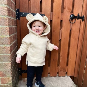 May include: A toddler wearing a beige hoodie with bear ears, black pants, and black and white sneakers. The child is smiling and standing in front of a wooden fence. The hoodie has a kangaroo pocket.