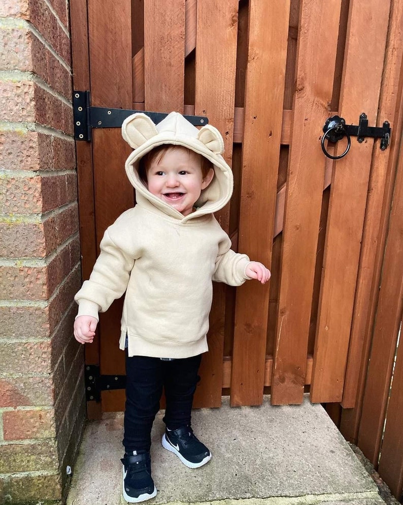 May include: A toddler wearing a beige hoodie with bear ears, black pants, and black and white sneakers. The child is smiling and standing in front of a wooden fence. The hoodie has a kangaroo pocket.