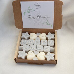 May include: A brown cardboard box with a white card that says "Happy Christmas" inside. The box is filled with small, white wax melts shaped like stars, hearts, and the letters "HAPPY XMAS".