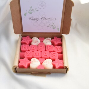 May include: A box of pink and white wax melts spelling out "Happy Xmas". The box is open and the wax melts are arranged in a pattern. The box has a card on top that says "Happy Christmas".