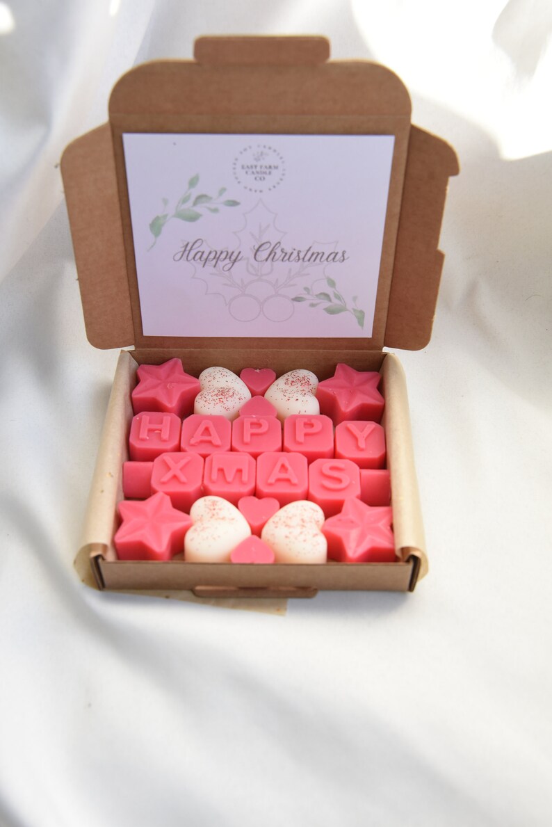 May include: A box of pink and white wax melts spelling out "Happy Xmas". The box is open and the wax melts are arranged in a pattern. The box has a card on top that says "Happy Christmas".