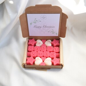 May include: A brown cardboard box with a pink and white card that says "Happy Christmas". The box is open and contains pink and white wax melts shaped like stars, hearts, and letters that spell out "HAPPY XMAS".