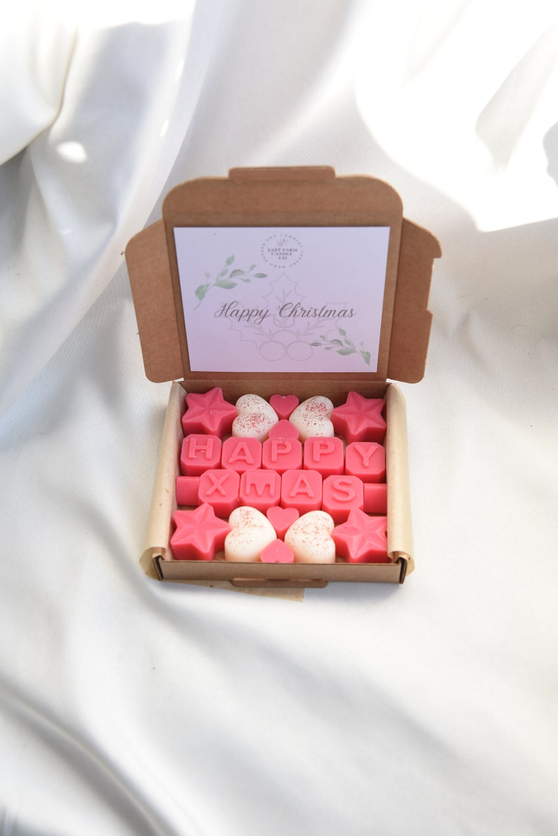 May include: A brown cardboard box with a pink and white card that says "Happy Christmas". The box is open and contains pink and white wax melts shaped like stars, hearts, and letters that spell out "HAPPY XMAS".