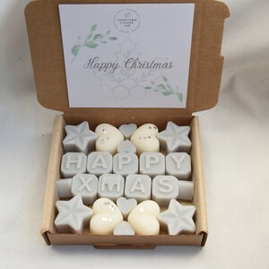 May include: A box of white wax melts with a "Happy Xmas" message. The melts are shaped like stars, hearts, and letters. The box is brown with a white card that says "Happy Christmas".