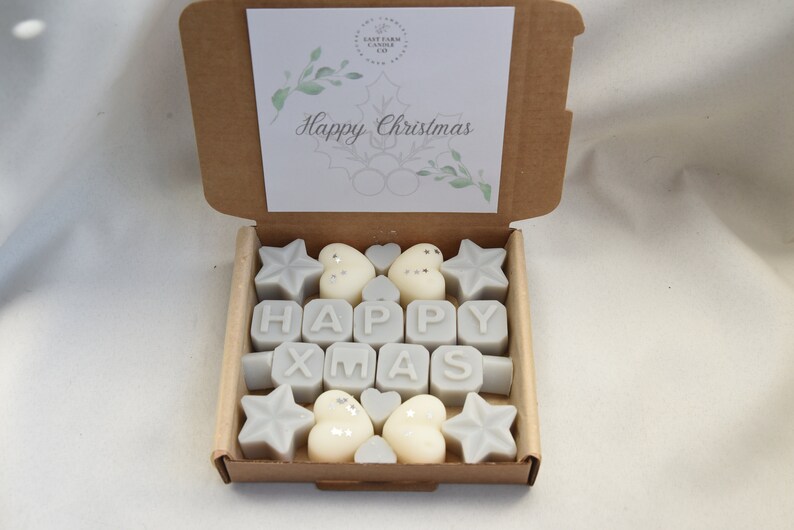 May include: A box of white wax melts with a "Happy Xmas" message. The melts are shaped like stars, hearts, and letters. The box is brown with a white card that says "Happy Christmas".