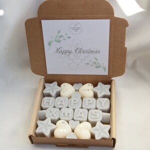 May include: A brown cardboard box with a white card that says "Happy Christmas" inside. The box is filled with small, white wax melts shaped like stars, hearts, and the letters "HAPPY XMAS".