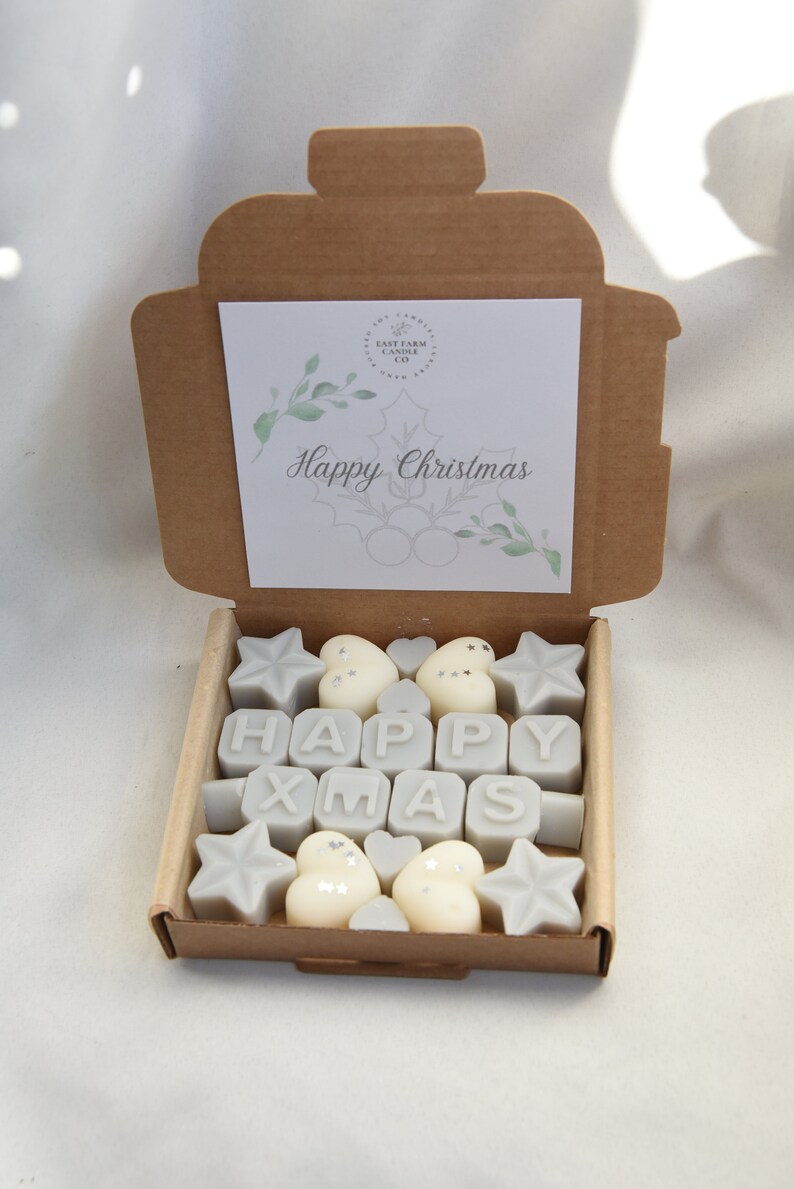 May include: A brown cardboard box with a white card that says "Happy Christmas" inside. The box is filled with small, white wax melts shaped like stars, hearts, and the letters "HAPPY XMAS".