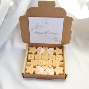 May include: A brown cardboard box with a lid open, revealing a set of wax melts in the shape of stars, hearts, and letters spelling "HAPPY XMAS". The box has a white card inside with the text "Happy Christmas" and a green floral design.