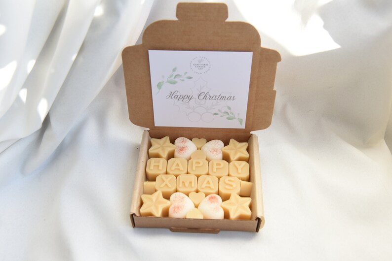May include: A brown cardboard box with a lid open, revealing a set of wax melts in the shape of stars, hearts, and letters spelling "HAPPY XMAS". The box has a white card inside with the text "Happy Christmas" and a green floral design.