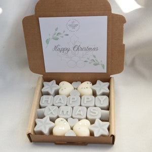 May include: A brown cardboard box with a white card inside that says "Happy Christmas". The box is open and contains a set of gray wax melts shaped like stars, hearts, and the letters "HAPPY XMAS".