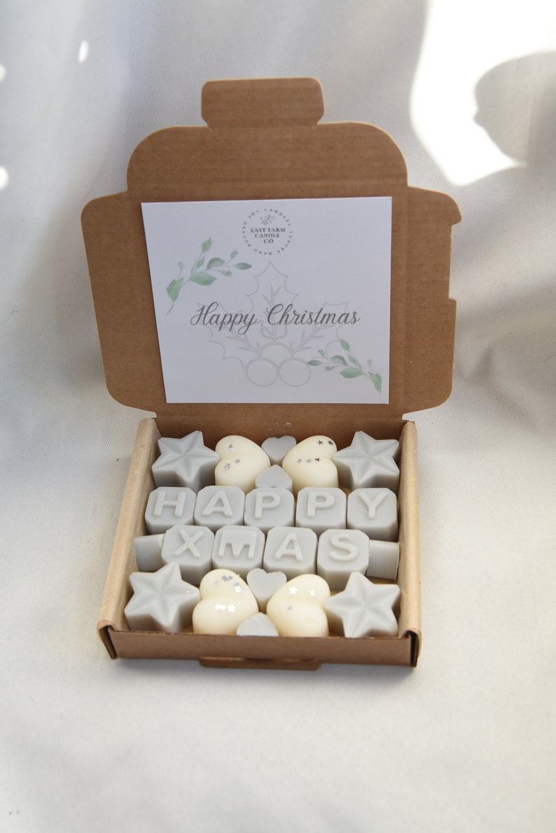 May include: A brown cardboard box with a white card inside that says "Happy Christmas". The box is open and contains a set of gray wax melts shaped like stars, hearts, and the letters "HAPPY XMAS".
