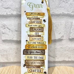 May include: A decorative bookmark with a gold tassel. The bookmark features the word "Gran" and a stack of book-shaped blocks with inspirational text about life's chapters. Small bee illustrations and floral accents are also present.