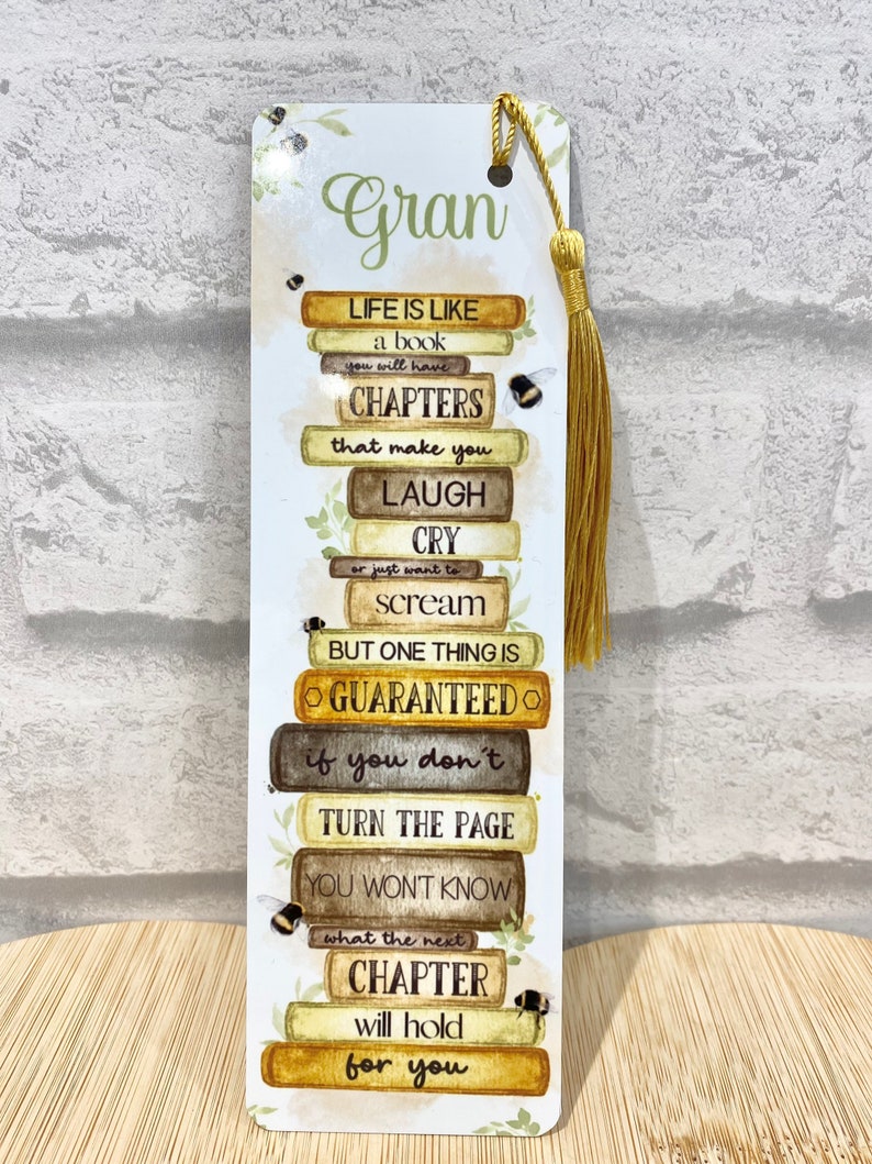 May include: A decorative bookmark with a gold tassel. The bookmark features the word "Gran" and a stack of book-shaped blocks with inspirational text about life's chapters. Small bee illustrations and floral accents are also present.