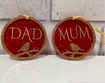 Pode incluir: Dois ornamentos circulares vermelhos com bordas de glitter dourado e fitas douradas para pendurar. Cada ornamento apresenta um pássaro dourado em um galho e o texto DAD ou MUM em letras douradas. A superfície vermelha tem textura marmorizada.