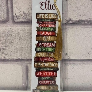 May include: A bookmark with a gold tassel, featuring a stack of books with the text "Life is like a book..." and the name "Ellie" in a decorative font. The bookmark has a white background.