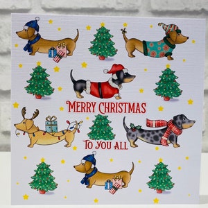 Pu&ograve; includere: Un biglietto di Natale con bassotti dei cartoni animati vestiti per le feste. I cani indossano costumi da Babbo Natale, sciarpe e cappelli, con alberi di Natale e stelle. Il testo "Merry Christmas To You All" &egrave; scritto in rosso.