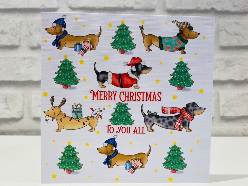 Pu&ograve; includere: Un biglietto di Natale con bassotti dei cartoni animati vestiti per le feste. I cani indossano costumi da Babbo Natale, sciarpe e cappelli, con alberi di Natale e stelle. Il testo "Merry Christmas To You All" &egrave; scritto in rosso.