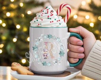 Pode incluir: Uma caneca de cerâmica branca com alça e borda em tom verde-azulado, apresentando um design estampado de uma letra R grande cercada por uma guirlanda de flocos de neve azuis e prateados, um pequeno quebra-nozes e uma caixa de presente.
