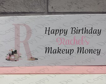 Pode incluir: Um cartão retangular branco com uma letra R grande em rosa pálido no lado esquerdo, decorada com pequenas ilustrações de maquilhagem. O texto diz Happy Birthday Rachel's Makeup Money em preto e rosa. O cartão repousa sobre um envelope rosa pálido.