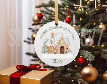 Pode incluir: Um enfeite de Natal redondo com um design de casa de gengibre. O enfeite tem o texto "OUR FIRST CHRISTMAS IN OUR NEW HOME" e o ano "2025". Ele pende de uma fita dourada. O fundo inclui uma árvore de Natal e um presente embrulhado.