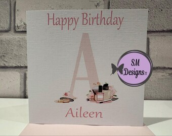 Pode incluir: Cartão de aniversário quadrado com as palavras "Happy Birthday" e "Aileen" em rosa. Uma grande letra "A" rosa no centro, decorada com maquiagem e flores. Um círculo roxo com uma borboleta e "SM Designs" está à direita.