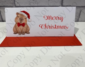 Pode incluir: Um cartão de felicitações retangular branco com uma capivara castanha usando um gorro de Pai Natal vermelho e uma gravata borboleta vermelha. O texto Merry Christmas está impresso a vermelho à direita. Um envelope vermelho está por baixo.