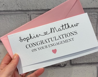 Pode incluir: Um cartão de noivado branco com os nomes "Sophie" e "Matthew" em itálico, e as palavras "CONGRATULATIONS ON YOUR ENGAGEMENT" em negrito. Um coração rosa está abaixo do texto. O cartão está dentro de um envelope rosa.