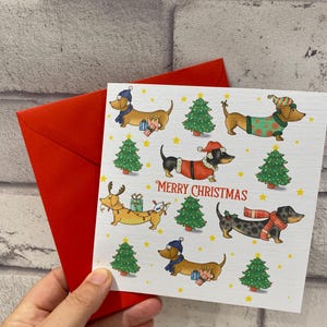 Pu&ograve; includere: Un biglietto di Natale con illustrazioni di bassotti in abiti festivi, come cappelli e sciarpe, insieme ad alberi di Natale e la scritta "Merry Christmas". Il biglietto &egrave; tenuto con una busta rossa.