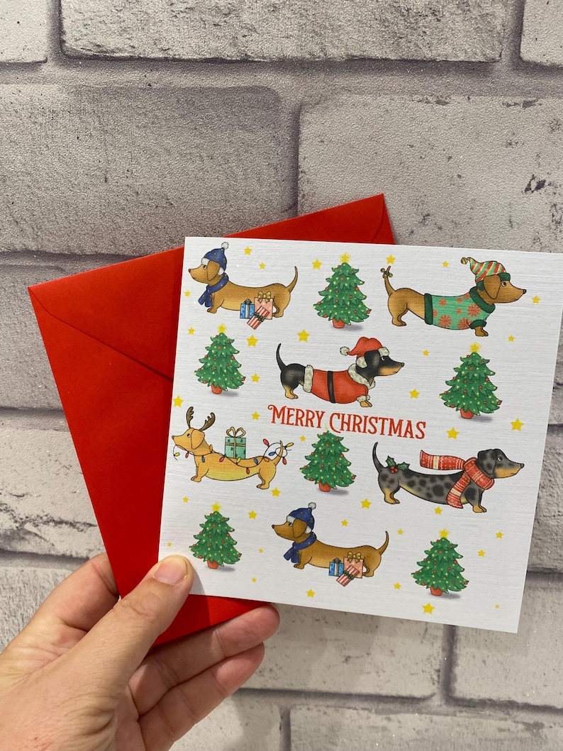 Pu&ograve; includere: Un biglietto di Natale con illustrazioni di bassotti in abiti festivi, come cappelli e sciarpe, insieme ad alberi di Natale e la scritta "Merry Christmas". Il biglietto &egrave; tenuto con una busta rossa.
