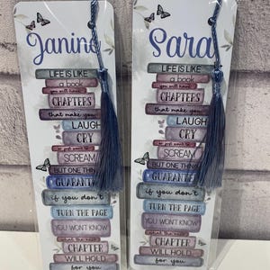 May include: Two personalized bookmarks with blue tassels. Each bookmark features a stack of books with the text "Life is like a book" and a quote about chapters. One bookmark says "Janine" and the other says "Sara".