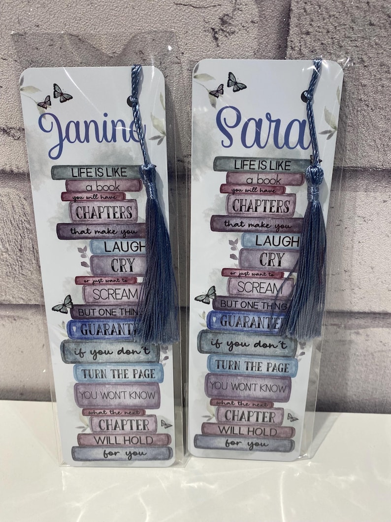 May include: Two personalized bookmarks with blue tassels. Each bookmark features a stack of books with the text "Life is like a book" and a quote about chapters. One bookmark says "Janine" and the other says "Sara".