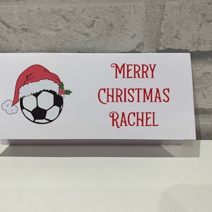 Può includere: Carta bianca con un pallone da calcio rosso e bianco che indossa un cappello da Babbo Natale. La carta dice "Buon Natale Rachel" in testo rosso.